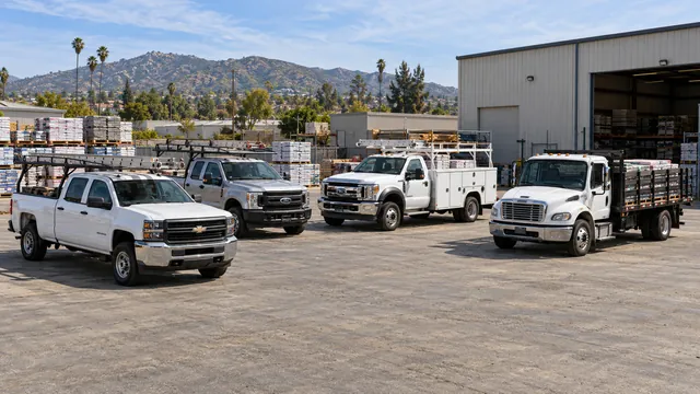 Work pickups with ladder racks, a service-body truck and a loaded stake bed truck at a building supply yard